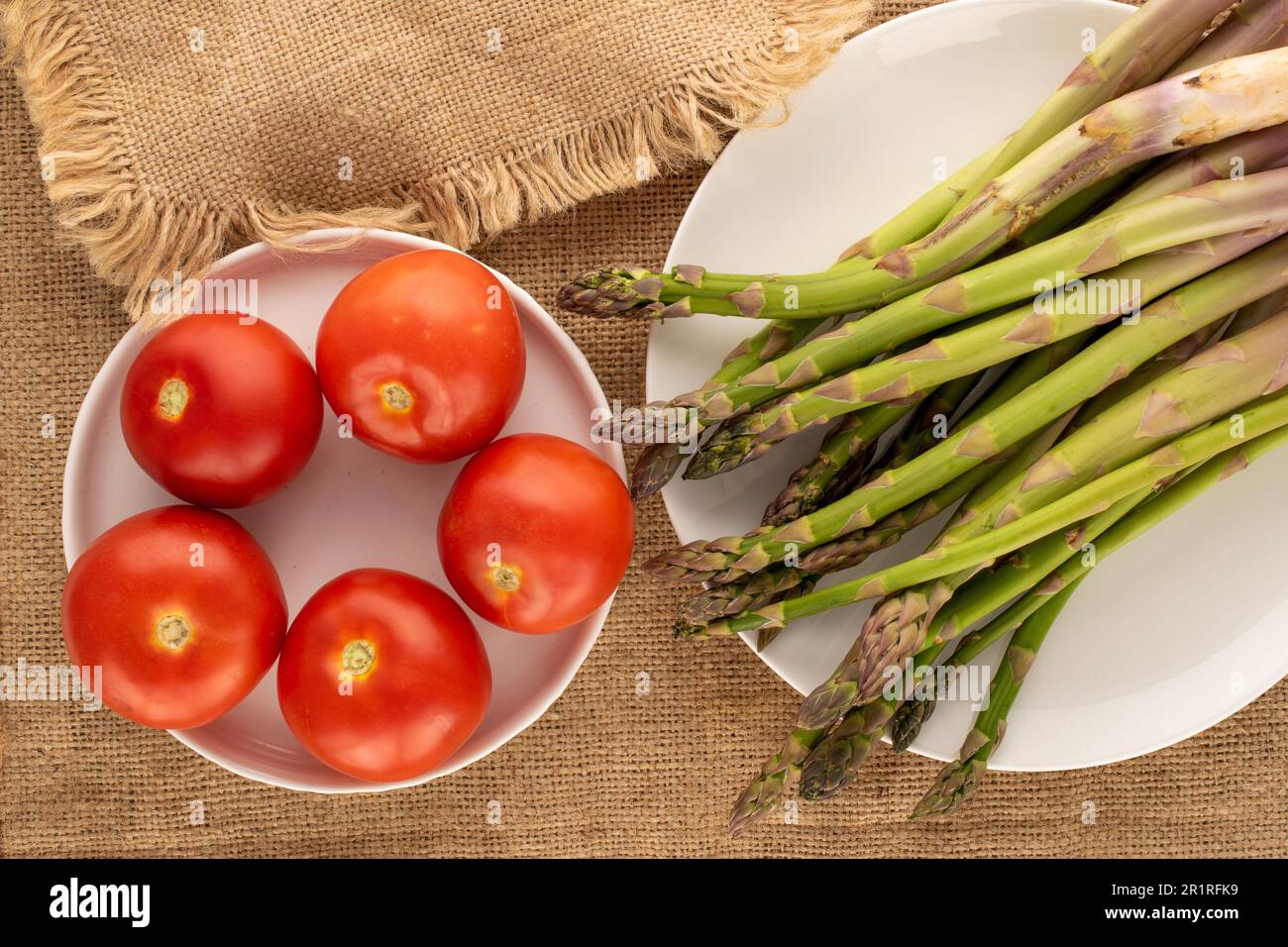 Mehrere Stiele organischen Spargels, fünf Tomaten mit weißen Keramikplatten auf Jutetuch, Makro, Draufsicht. Stockfoto
