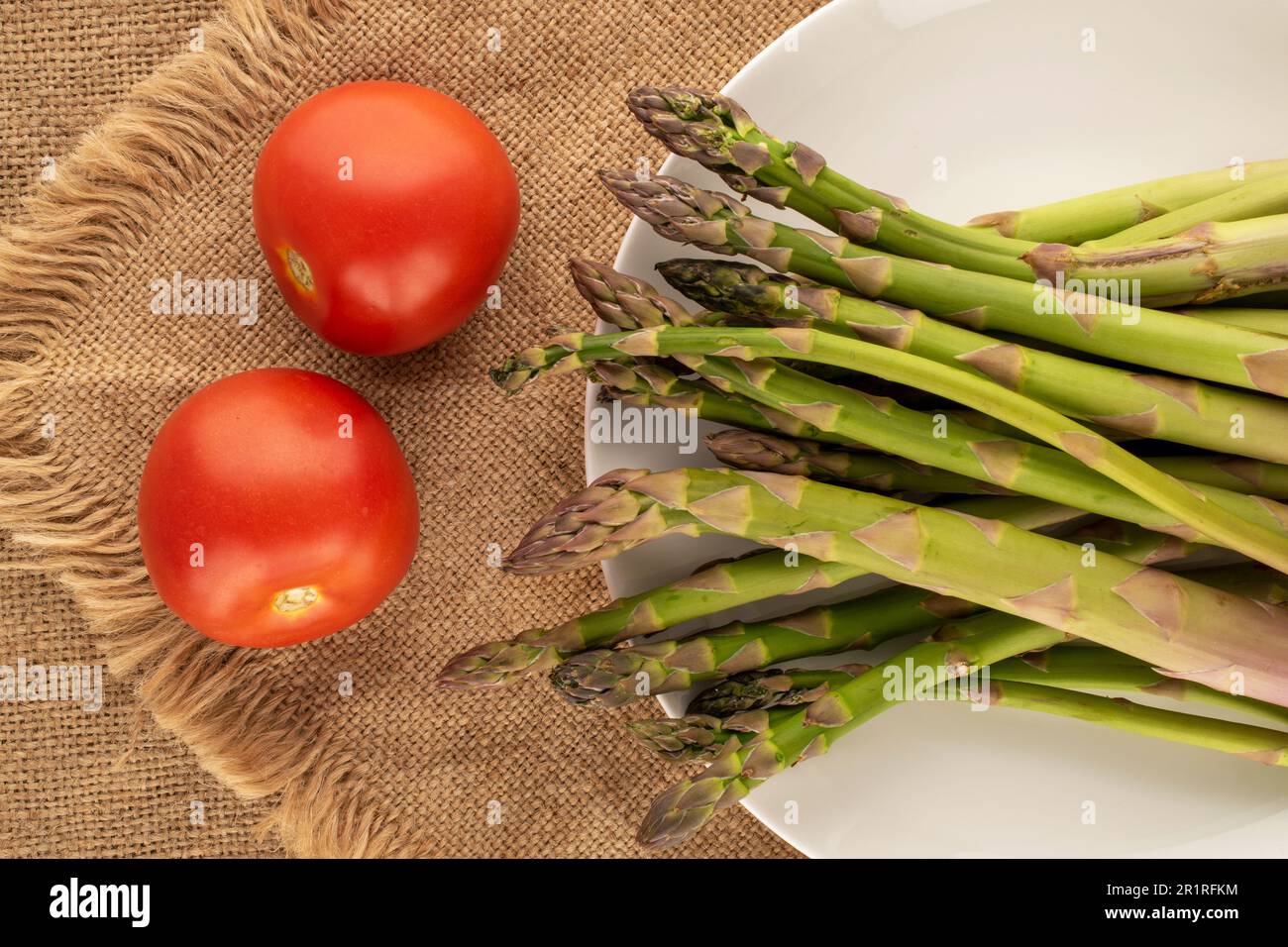 Mehrere Stiele organischen Spargels, zwei Tomaten mit weißer Keramikplatte auf Jutetuch, Makro, Draufsicht. Stockfoto