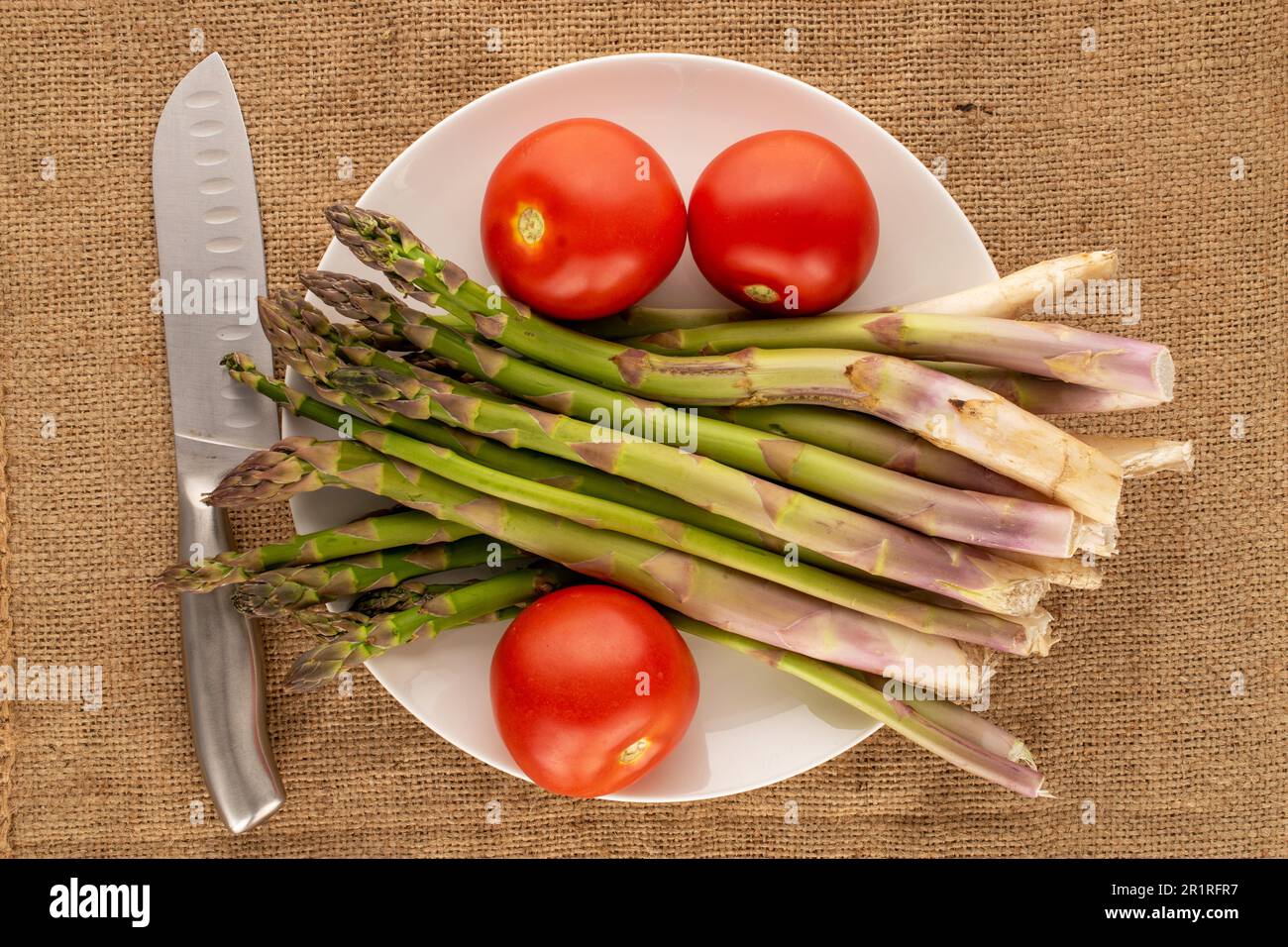 Mehrere Stiele organischen Spargels, drei Tomaten und ein Metallmesser mit weißer Keramikplatte auf Jutetuch, Makro, Draufsicht. Stockfoto