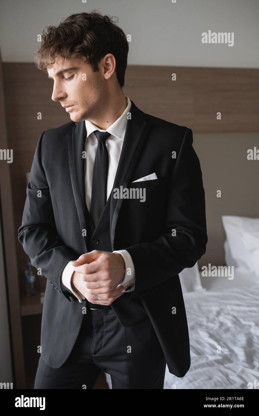 Hübscher Bräutigam in formeller Kleidung mit eleganter schwarzer Krawatte und weißem Hemd, der im modernen Hotelzimmer steht und die Handschellen anlegt, Mann am Hochzeitstag, bitte Stockfoto