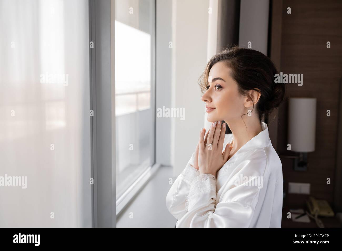 Seitenansicht einer hoffnungsvollen jungen Frau mit Verlobungsring am Finger, in weißem Seidenmantel mit betenden Händen und Blick durch das Fenster im Hotel sui Stockfoto