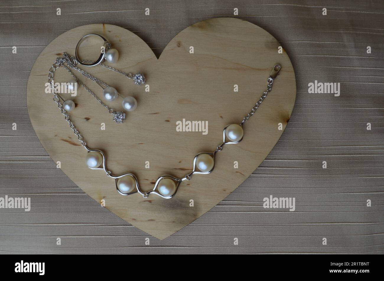 Ein hölzernes Herz zum Valentinstag mit silbernen Ornamenten, Ohrringen, Ringen, Halsketten mit Perlen, Diamanten, Edelsteine, Strasssteine auf einem beigen Ba Stockfoto