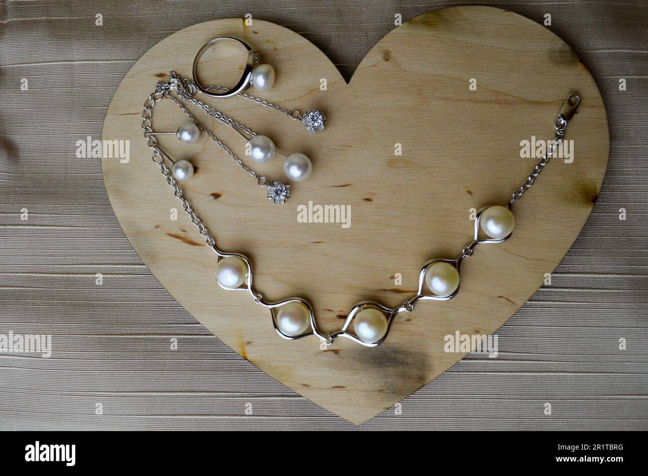 Ein hölzernes Herz zum Valentinstag mit silbernen Ornamenten, Ohrringen, Ringen, Halsketten mit Perlen, Diamanten, Edelsteine, Strasssteine auf einem beigen Ba Stockfoto