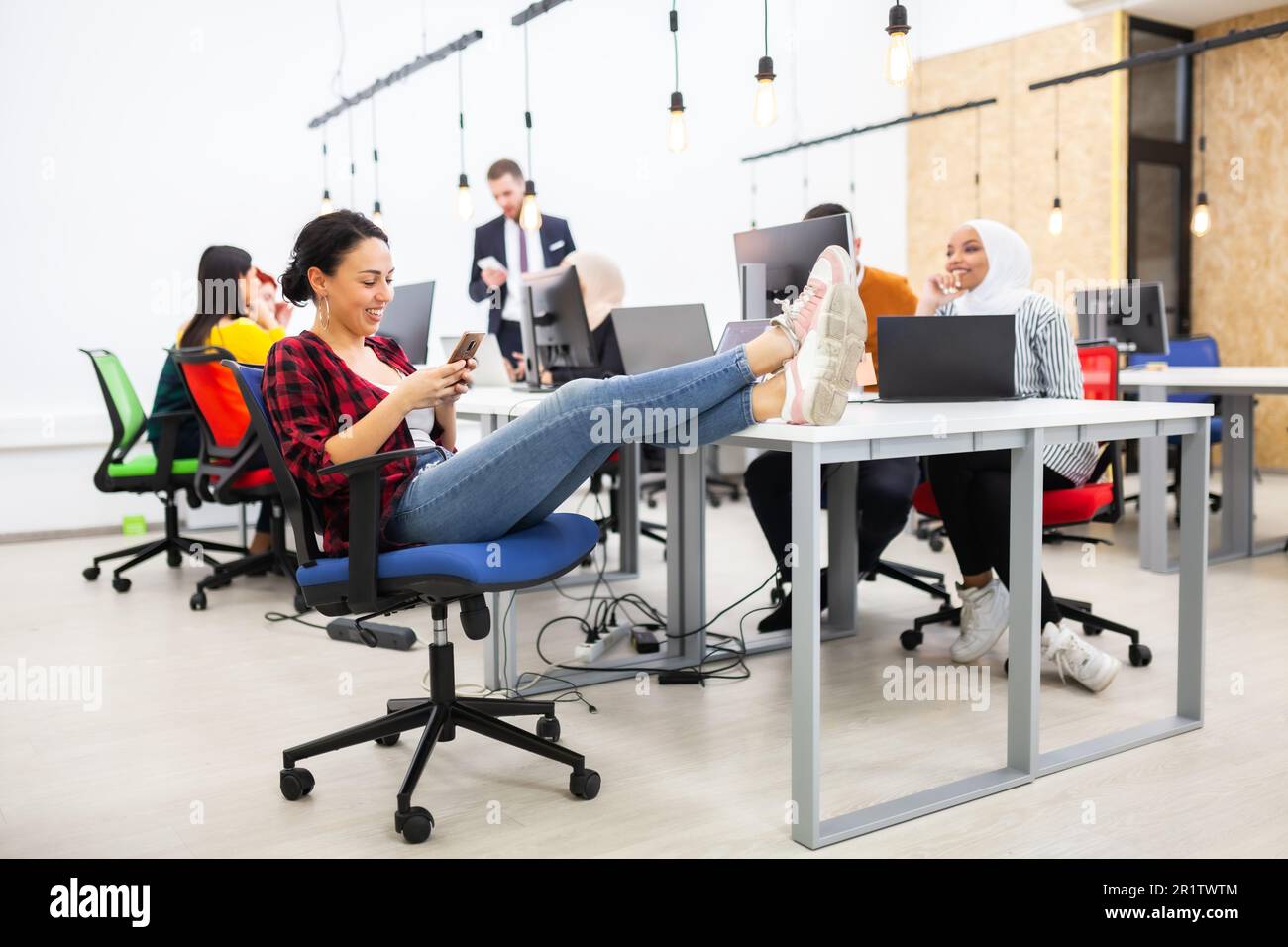 Gruppe von multiethnischen Kollegen, die an Desktop-Computern in einem modernen Bürobereich arbeiten. Mädchen ruht sich mit den Füßen auf dem Tisch aus Stockfoto