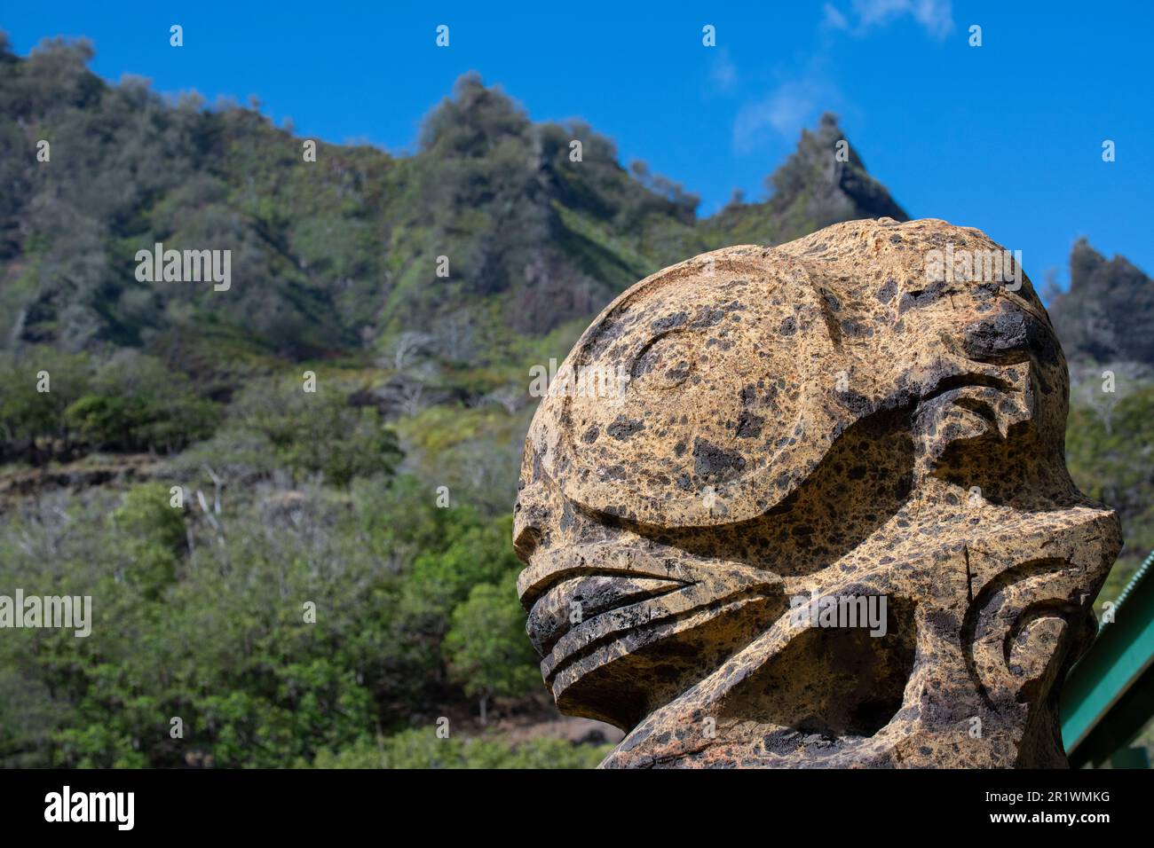 Französisch-Polynesien, Süd-Marquesas, Fatu Hiva, Dorf Omoa. Traditionelle Steinschnitzerei mit steilen vulkanischen Bergen in der Ferne. Stockfoto