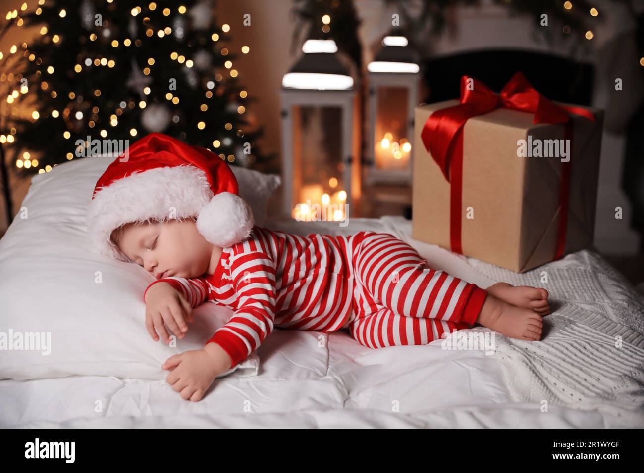 Baby im Weihnachtsschlafanzug und Weihnachtsmannmütze schlafen in der Nähe der Geschenkbox auf dem Bett drinnen Stockfoto