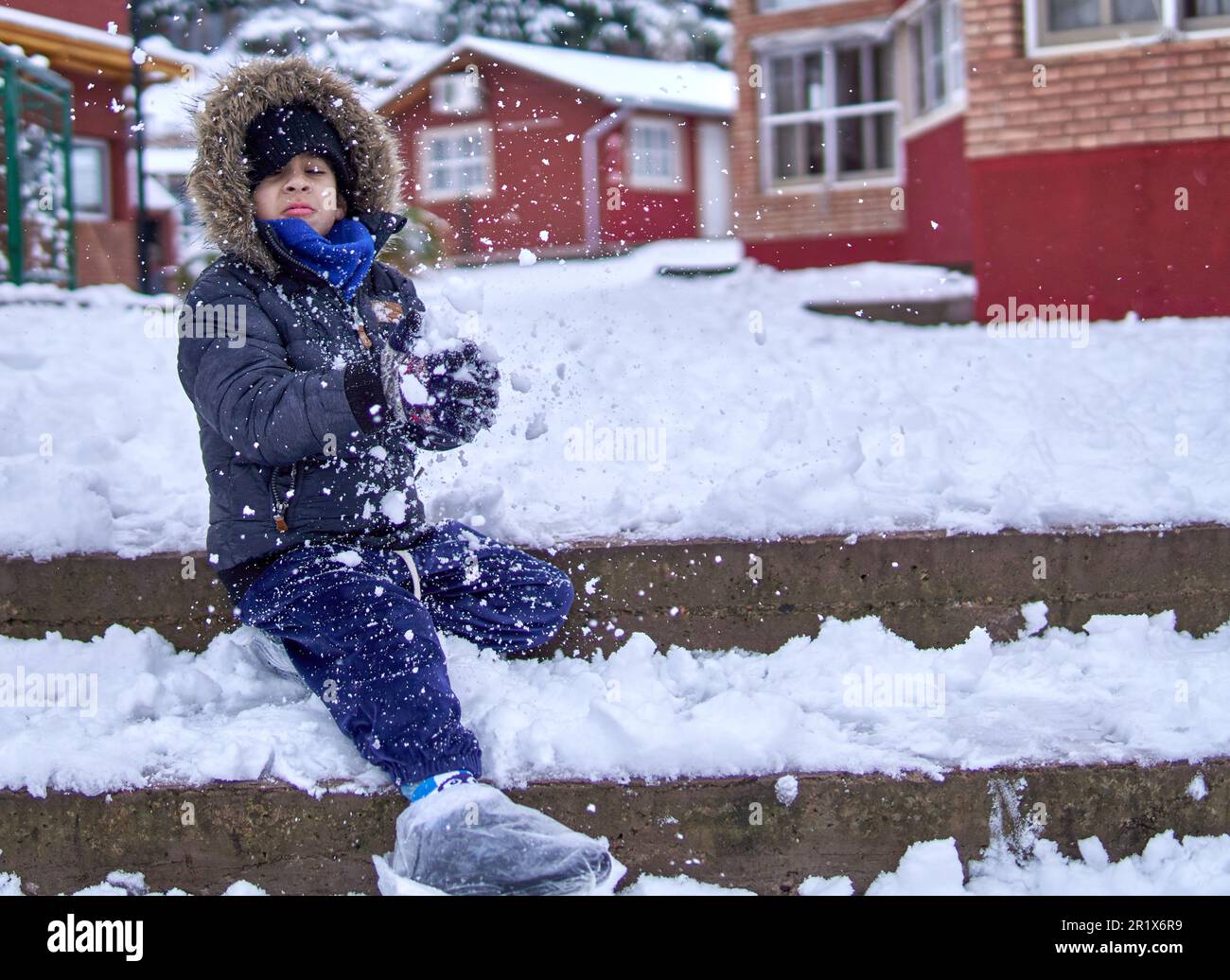 Lateinisches Kind, das auf einer Leiter vor einem Hüttenkomplex sitzt und mit Schnee spielt, einen warmen Parka in einer Pelzhaube trägt und die Schuhe mit Taschen bedeckt. Spritzen und Selen Stockfoto