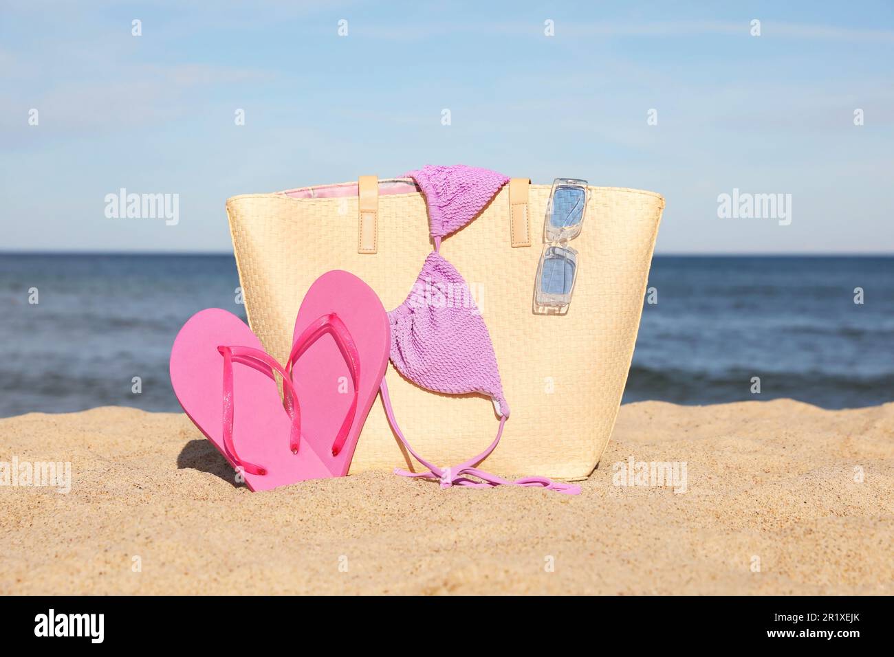 Sommertasche mit Hausschuhen, Bikinioberteil und Sonnenbrille auf Sand nahe dem Meer Stockfoto