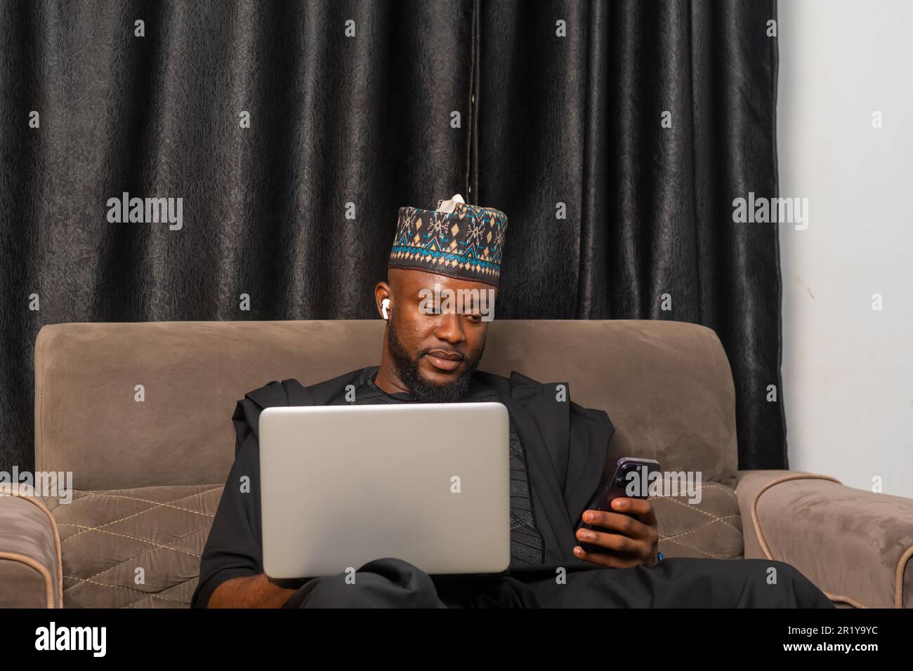 Gut aussehender Geschäftsmann in afrikanischer Kleidung, der an seinem Laptop arbeitete und sein Handy für geschäftliche Transaktionen benutzte Stockfoto