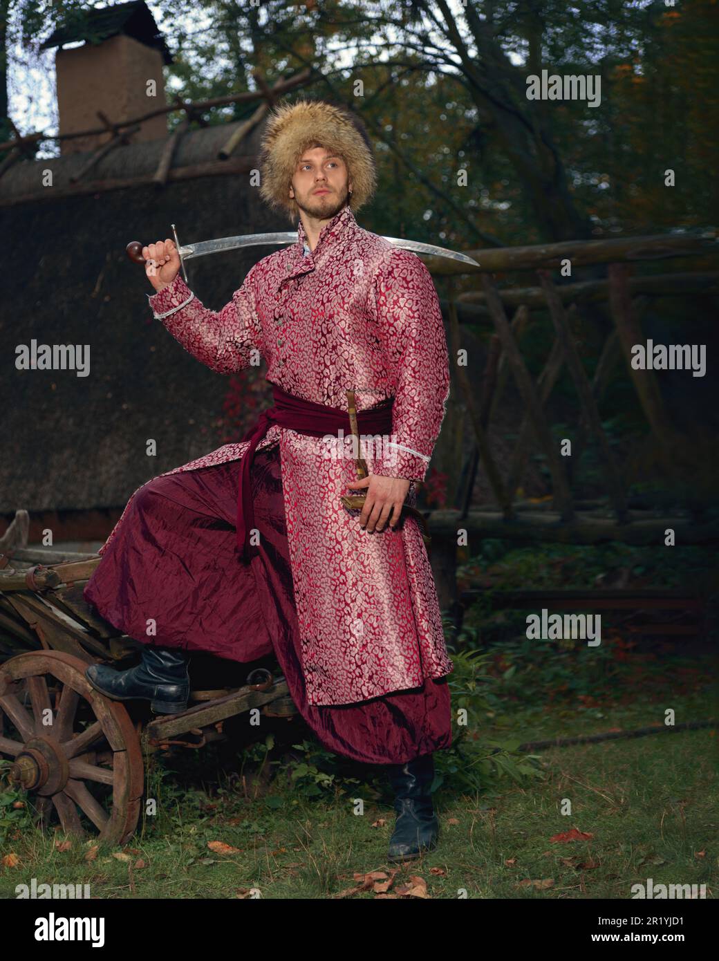 Selbstbewusster, tapferer junger ukrainischer kosack gekleidet in kunstvoller, traditioneller Kleidung und Pelzmütze, die den Säbel auf der Schulter hält und wegschaut Stockfoto