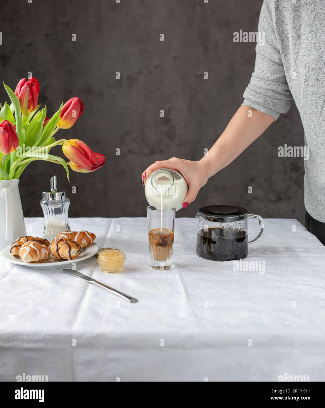 Frauenhände gießen Milch in den Kaffee. Das Frühstück steht auf dem Tisch. Stockfoto