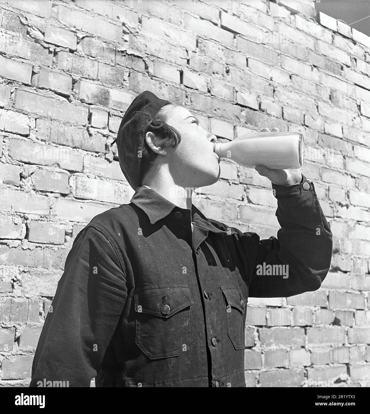 In den 1950er Jahren. Eine junge Lehrling, Maurerin, trinkt während einer Arbeitspause aus einer Milchflasche in der Sonne. Im Hintergrund ist eine neu errichtete Ziegelwand zu sehen. Zu dieser Zeit wurde Milch verkauft und in Glasflaschen geliefert oder man konnte Milch kaufen, die man aus dem Geschäft in seiner eigenen Flasche mitbringen konnte. Schweden 1951. Kristoffersson Ref. BB94-11 Stockfoto