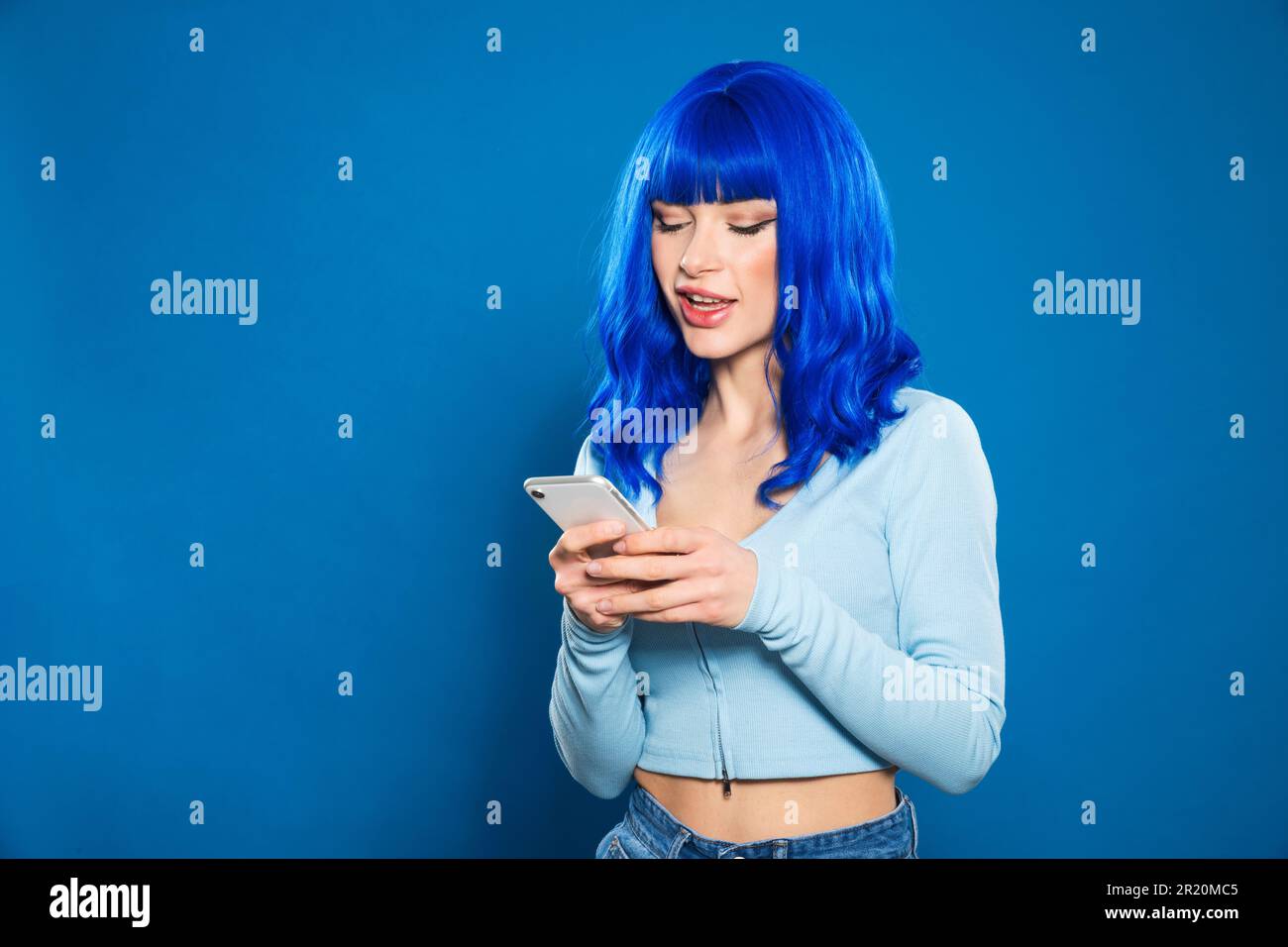 Positive junge Frau mit gefärbtem blauem Haar in legerer Kleidung, die vor blauem Hintergrund auf dem Handy surft Stockfoto