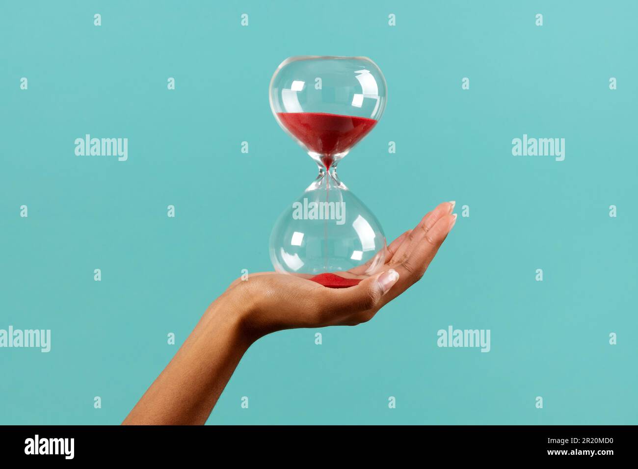 Nicht erkennbare schwarze weibliche Hand mit transparenter Sanduhr mit rotem Sand vor türkisfarbenem Hintergrund Stockfoto