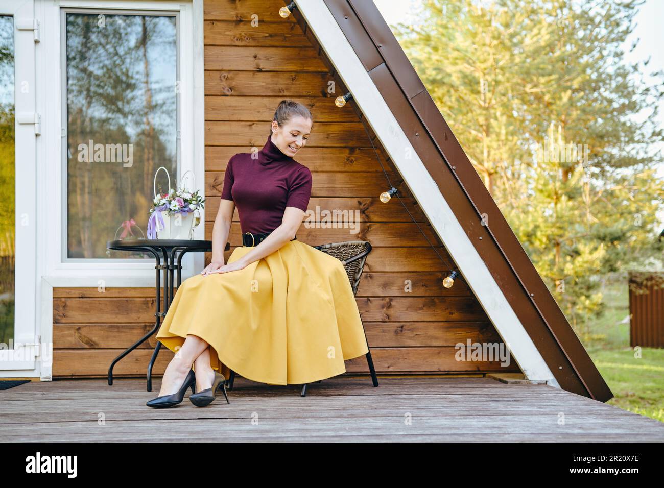 Die süße schüchterne Frau in einem langen Rock und einer Bluse sitzt auf der Terrasse der Holzkabine Stockfoto