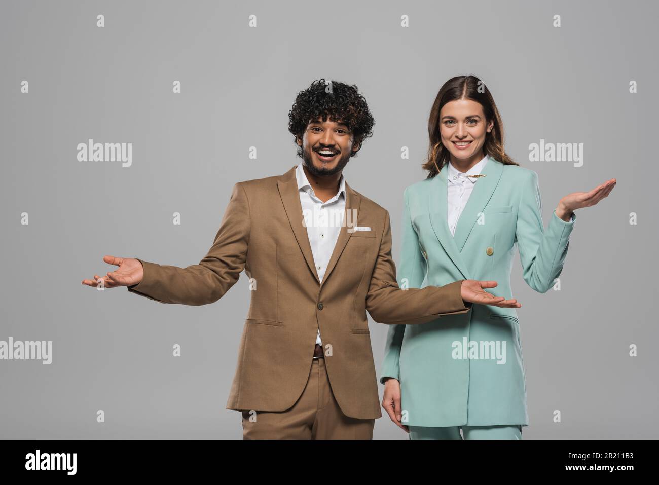 Sorgenfreies multiethnisches Event Gastgeber in verschiedenen Farben formelle Kleidung zeigt mit Händen zusammen und schaut während des Urlaubs isoliert auf grau, St. Stockfoto