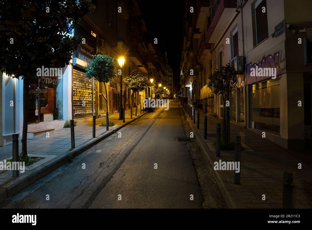 Thessaloniki, Griechenland, 27. April 2023: Dunkle, enge Straße bei Nacht mit älteren Wohnhäusern, Geschäften und kleinen Bäumen zwischen den Laternen in der Cit Stockfoto