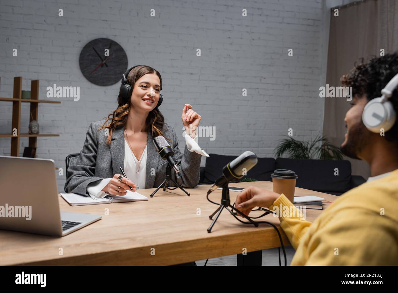 Positive weibliche Podcast-Moderatorin in kabellosen Kopfhörern, die mit verschwommenem indischen Gast spricht, während sie mit einem Stift neben Notebook, Geräten, Mikrofon und Coff sitzt Stockfoto