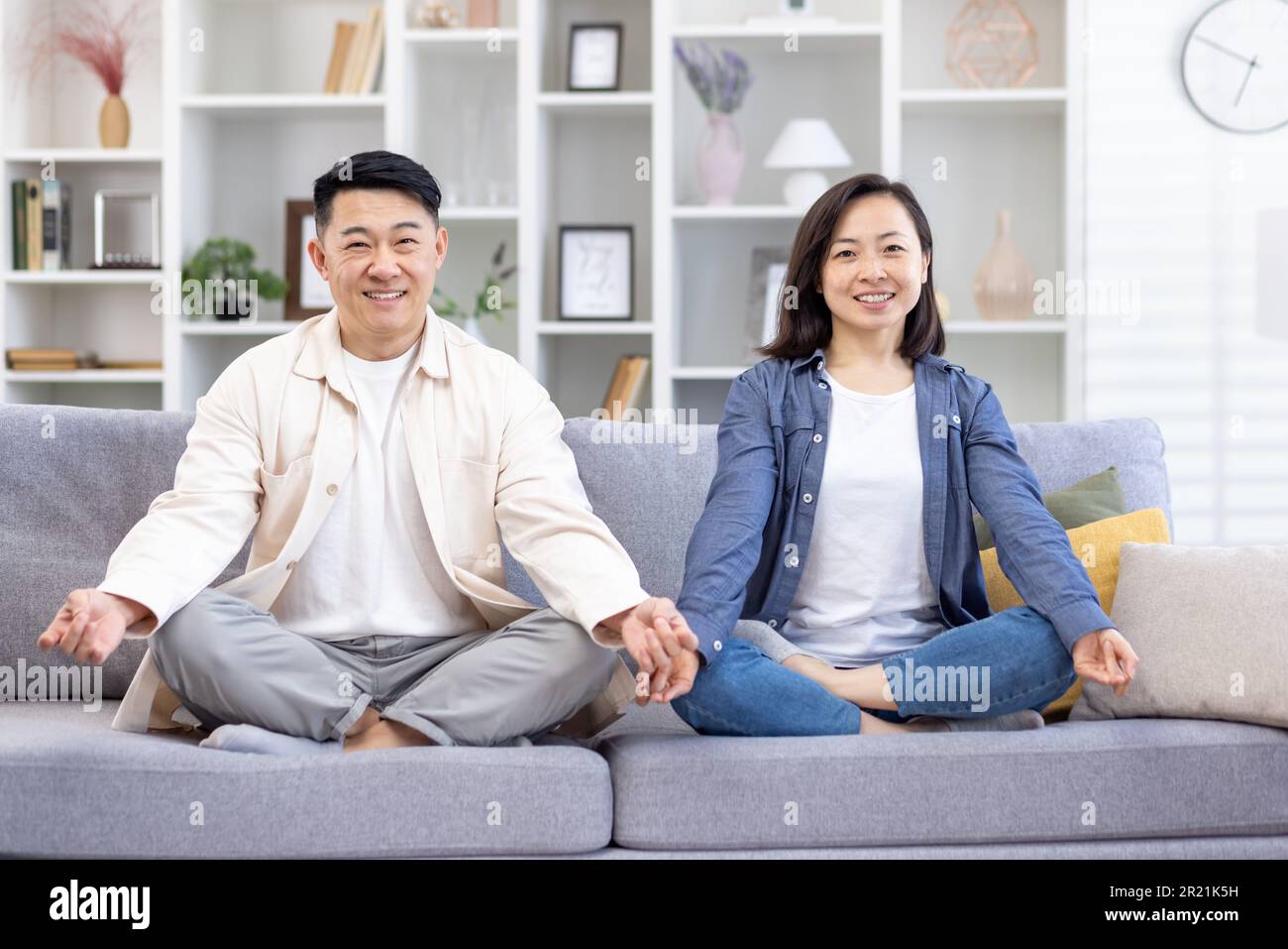 Porträt einer jungen asiatischen Familie, ein Paar, das zu Hause Yoga macht. Ein Mann und eine Frau sitzen auf einem Sofa in Lotusposition und lächeln in die Kamera. Kümmern Sie sich um ihre psychische Gesundheit. Stockfoto