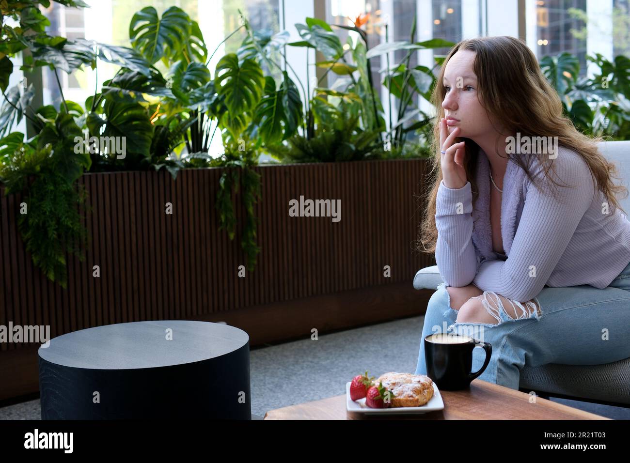 Ein junges Mädchen in zerrissenen Jeans sitzt in einem Café trauriges, reiches Leben ist nicht immer glücklich Traurigkeit melancholische Einsamkeit köstliches Essen Kaffee trinken allein Adoleszenz psychologische Probleme der blonden Kindheit Stockfoto