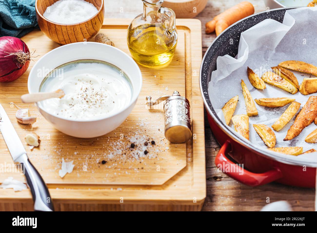 Backpfanne gefüllt mit Roastkartoffeln oder gebackenen Pommes frites in einer chaotischen Arbeitsküche mit einer Schüssel Joghurt-Sauce, Olivenöl, Knoblauch und Gewürzen und Con Stockfoto