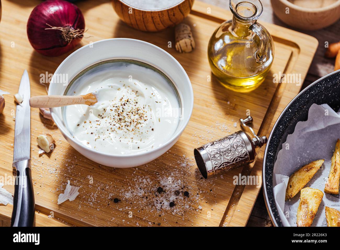 Backpfanne gefüllt mit Roastkartoffeln oder gebackenen Pommes frites in einer chaotischen Arbeitsküche mit einer Schüssel Joghurt-Sauce, Olivenöl, Knoblauch und Gewürzen und Con Stockfoto