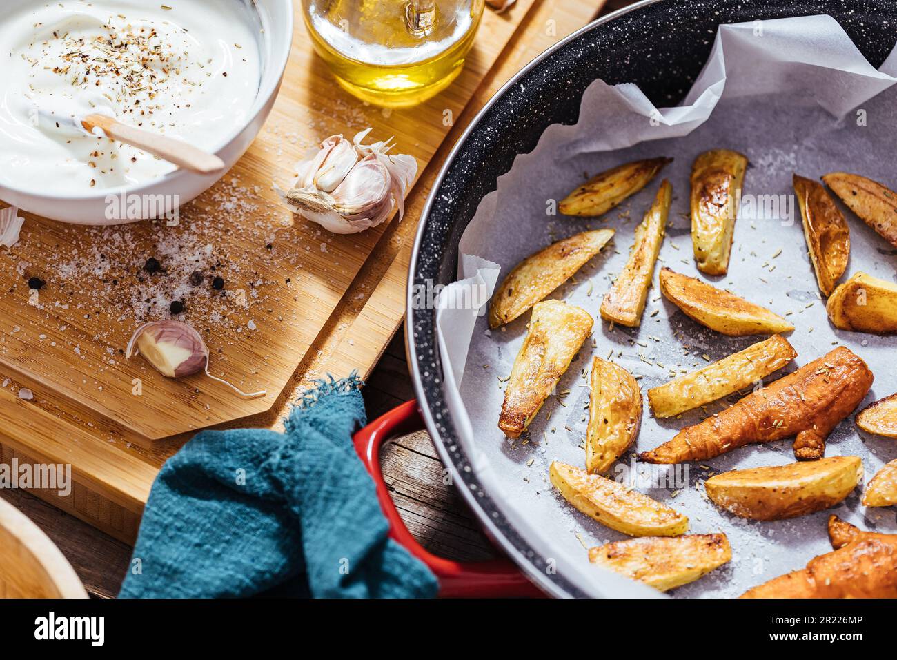 Backpfanne gefüllt mit Roastkartoffeln oder gebackenen Pommes frites in einer chaotischen Arbeitsküche mit einer Schüssel Joghurt-Sauce, Olivenöl, Knoblauch und Gewürzen und Con Stockfoto
