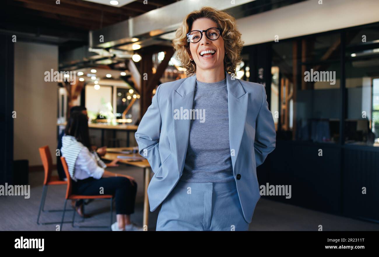 Frau, die in einem Büro steht, lächelt und trägt einen Anzug. Geschäftsfrau, die mit ihren Kollegen arbeitet, die im Hintergrund ein Meeting abhalten. Stockfoto