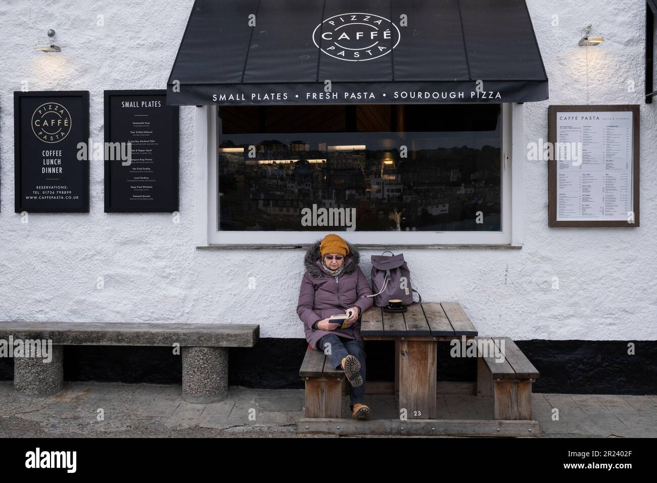 Eine Besucherin, die ein Buch liest und an einem kalten, kalten Tag in Cornwall in England außerhalb der Saison in St. Ives vor einem Restaurant sitzt. Stockfoto