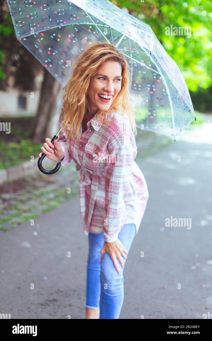 Fröhliche junge rothaarige Großstadtmodel-Frau mit langen welligen Haaren und transparentem Regenschirm, die draußen posiert Stockfoto