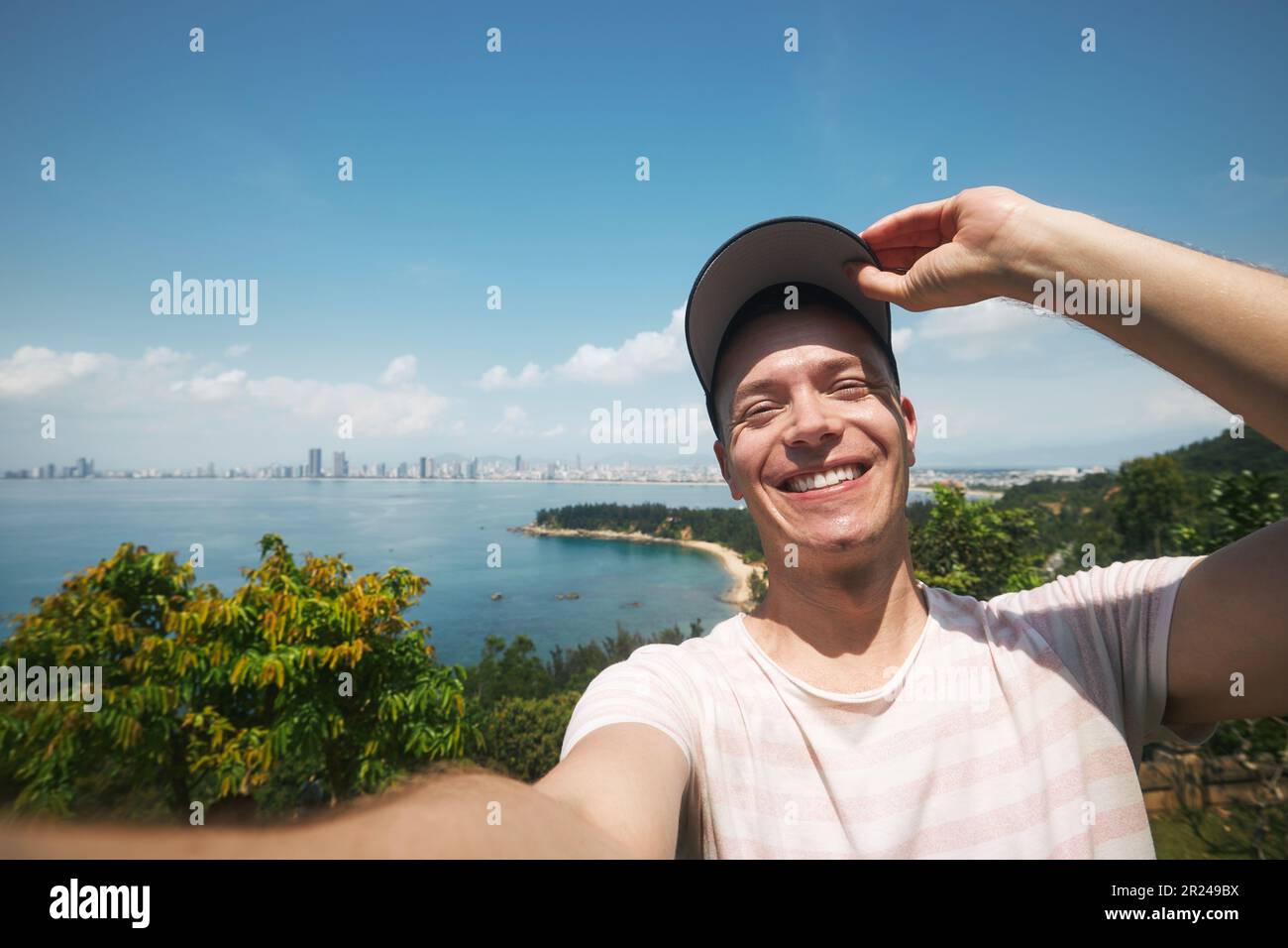 Glücklicher Mann, der ein Selfie-Foto vom Sommerferientag macht. Hübscher Tourist mit Mütze und Lächeln vor der Kamera vor der Landschaft mit Strand. Stockfoto
