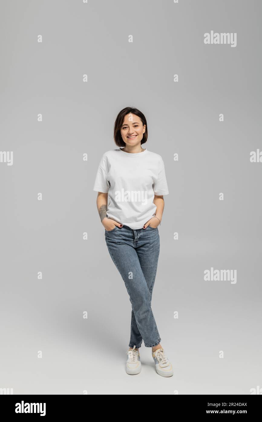 Lange, glückliche und tätowierte Frau mit kurzen Haaren und natürlichem Make-up, die in einem weißen T-Shirt steht und mit Händen in den Taschen des blauen Jeans posiert Stockfoto