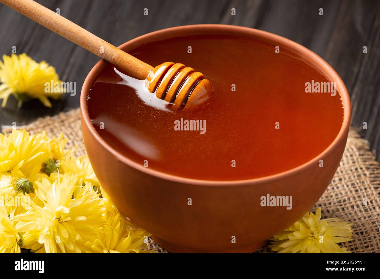 Honig in einer Tonschüssel, Dipper und gelbe Blumen auf Textil- und dunklem Holzhintergrund. Honigstäbchen liegen in einer Keramikschüssel mit Honignähe Stockfoto