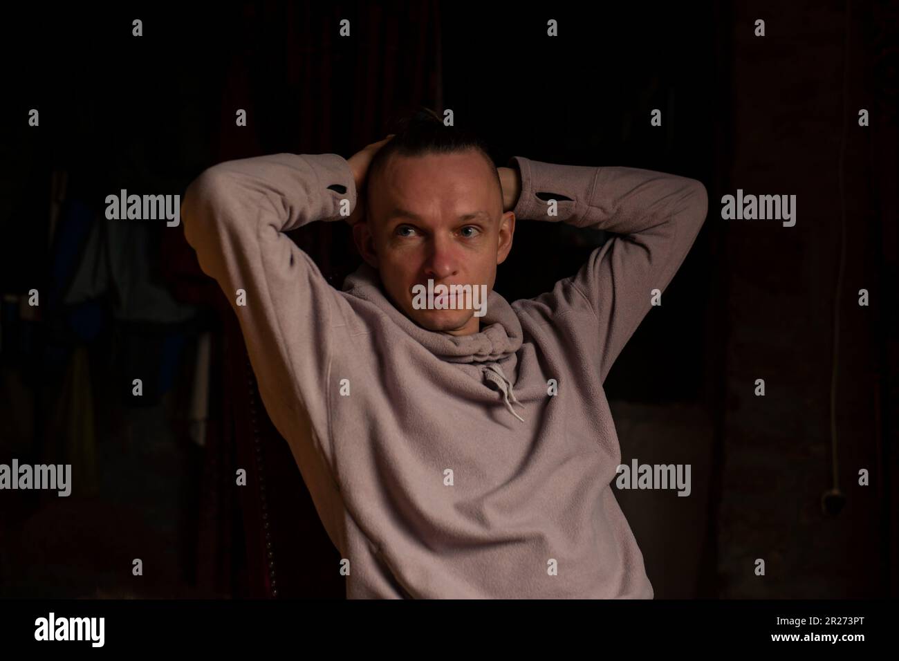 Ein unachtsamer Mann mittleren Alters in einem dunklen Raum. Entspannte Haltung, Hände hinter den Kopf. Dunkler Hintergrund, Freizeitkleidung. Stockfoto