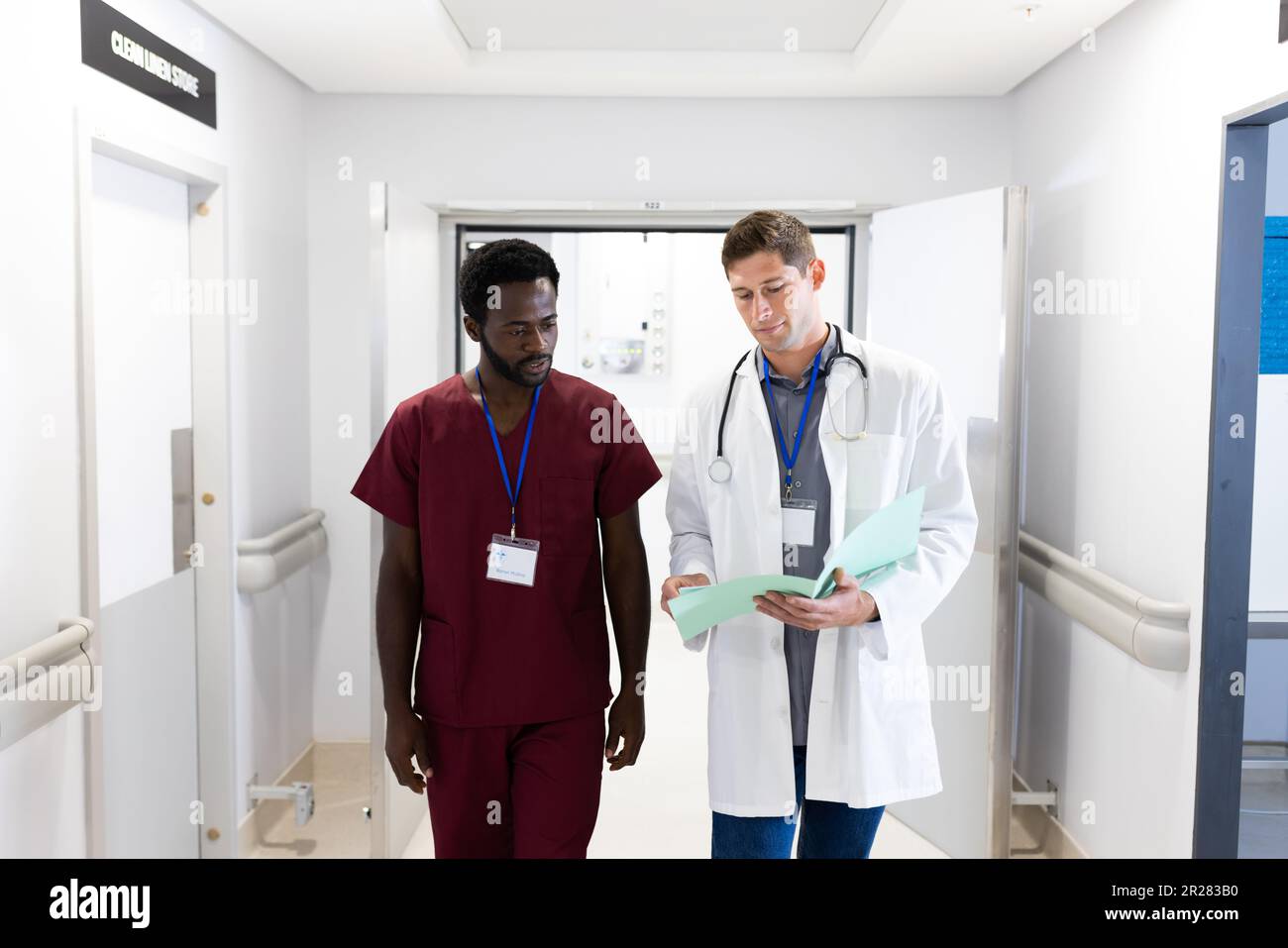 Zwei verschiedene männliche Ärzte schauen auf Notizen und gehen im Krankenhausflur Stockfoto
