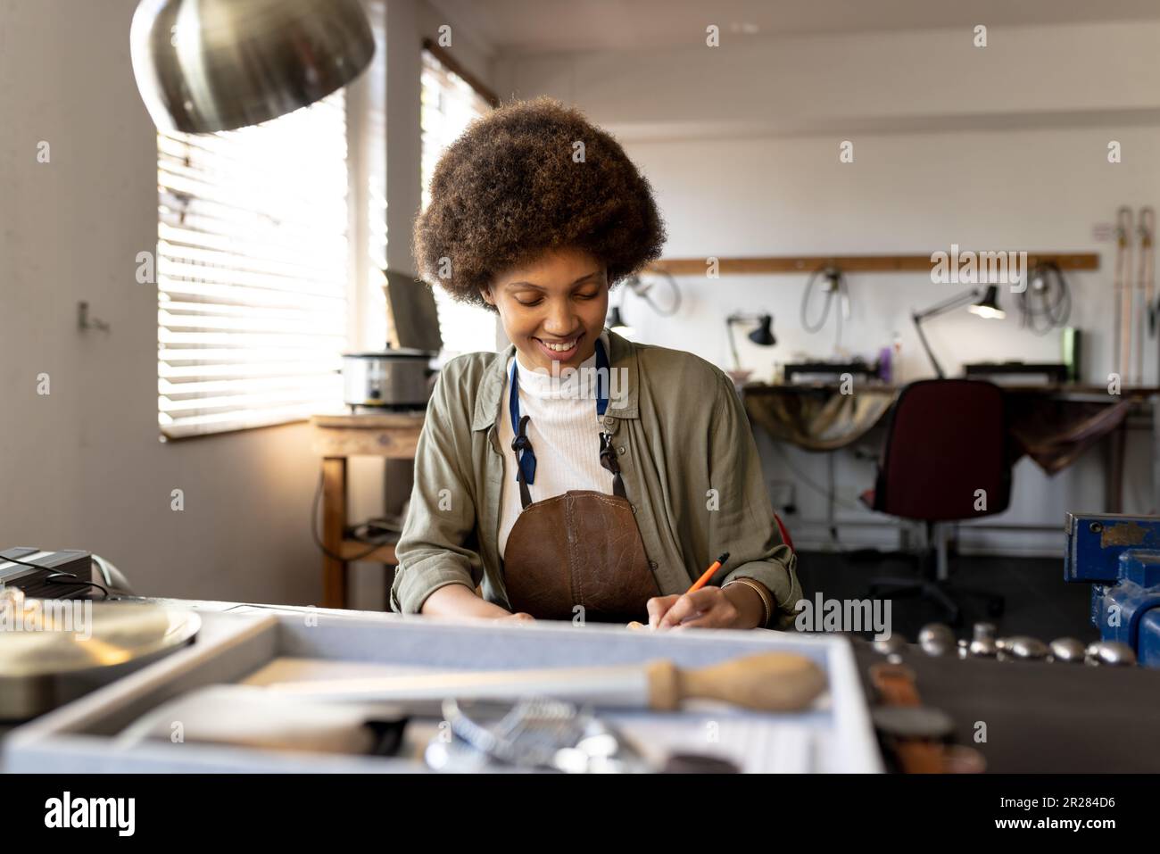 Glückliche birassische Schmuckdesignerin am Schreibtisch, die Designs in der Werkstatt skizziert Stockfoto