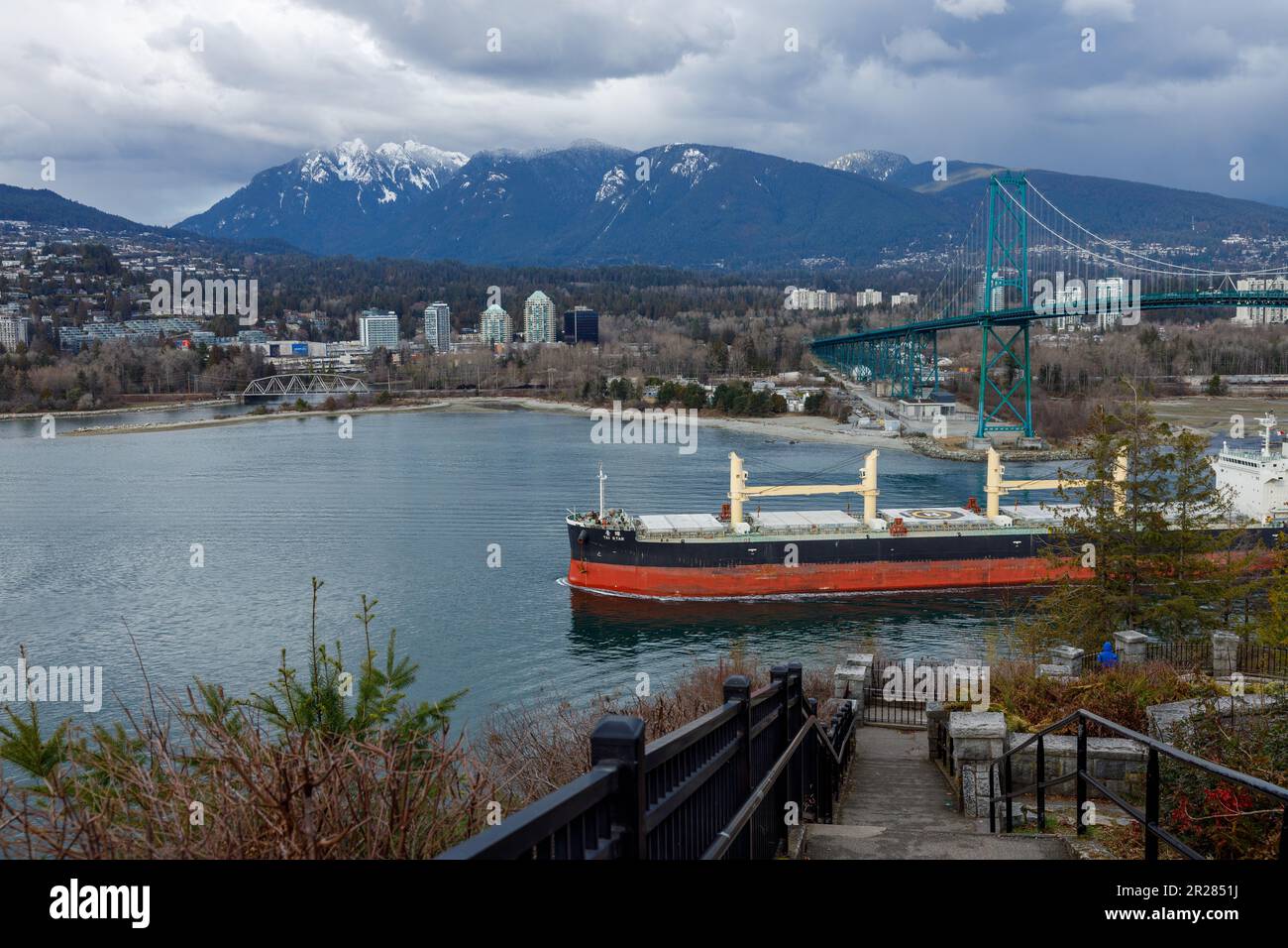 Vancouver, Kanada - März 8,2023: Ein riesiges Frachtschiff navigiert unter der Lions Gate Bridge in Burrard Inlet, mit der Skyline von West Vancouver hinten Stockfoto