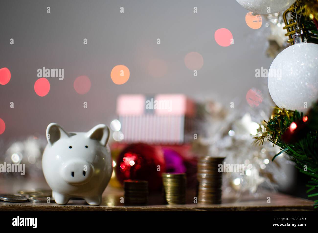 Nahaufnahme, weiße Sparschweine, viele Münzen auf Tischhölzern. Grüner Weihnachtsbaum mit mittelgroßen Kugeln. Konzeptinvestition Budget Wealth Business du Stockfoto