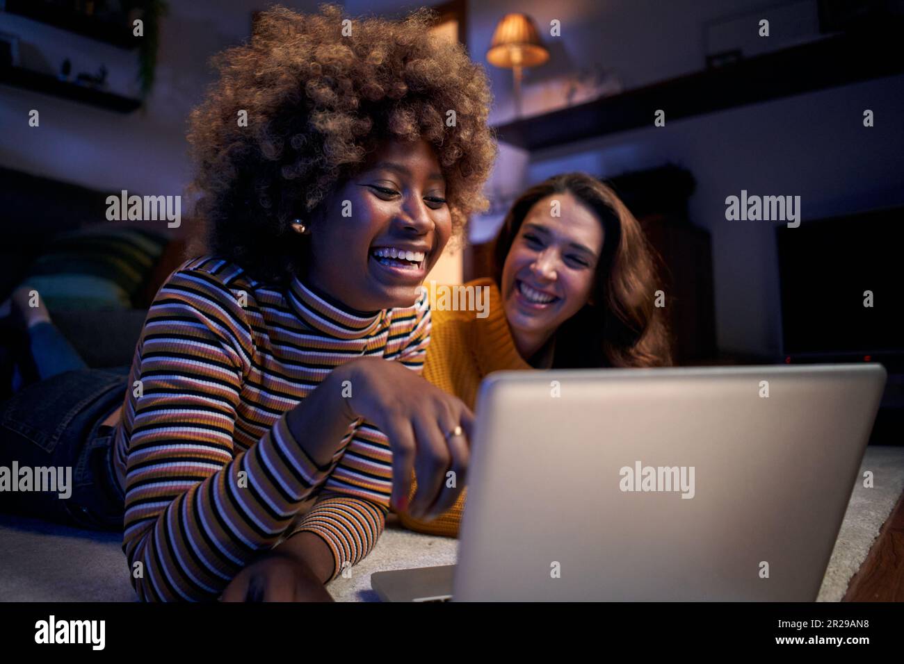 Zwei fröhliche, multiethnische Frauen, die auf einem Teppich in einem dunklen Wohnzimmer auf dem Computer liegen. Stockfoto