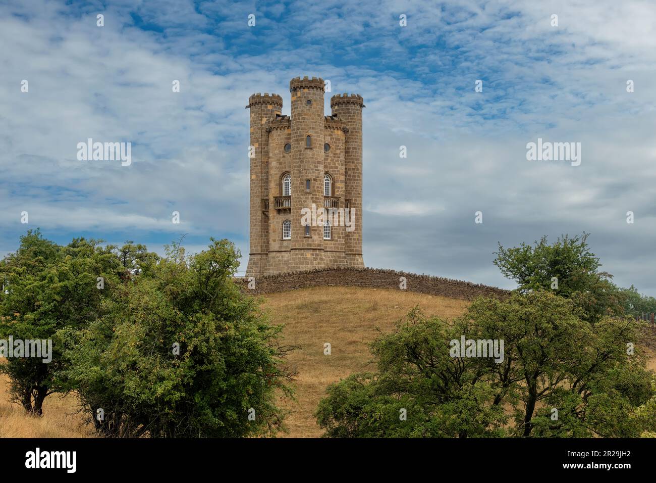 Niedriger Winkel Blick auf den Broadway Tower auf dem Beacon Hill entlang der Route des Cotswold Way in England mit Bäumen und Kalksteinwänden vor sich Stockfoto