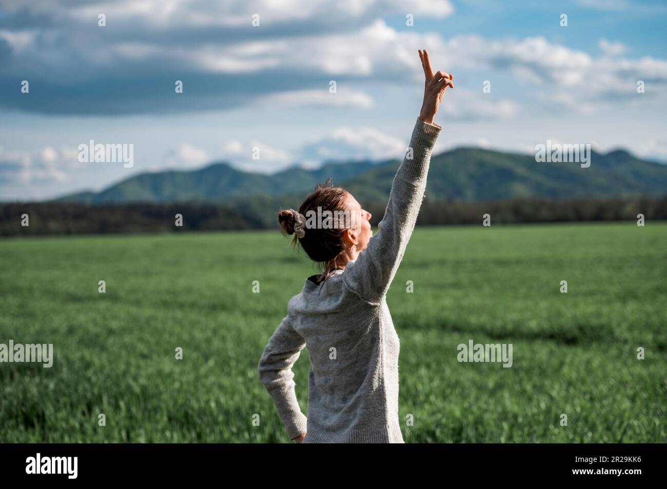 Authentische junge Frau, die unter dem dramatischen Himmel steht und ihren Arm in siegreicher Geste hochhebt. Wir feiern Leben und Erfolg umgeben von Grün Stockfoto