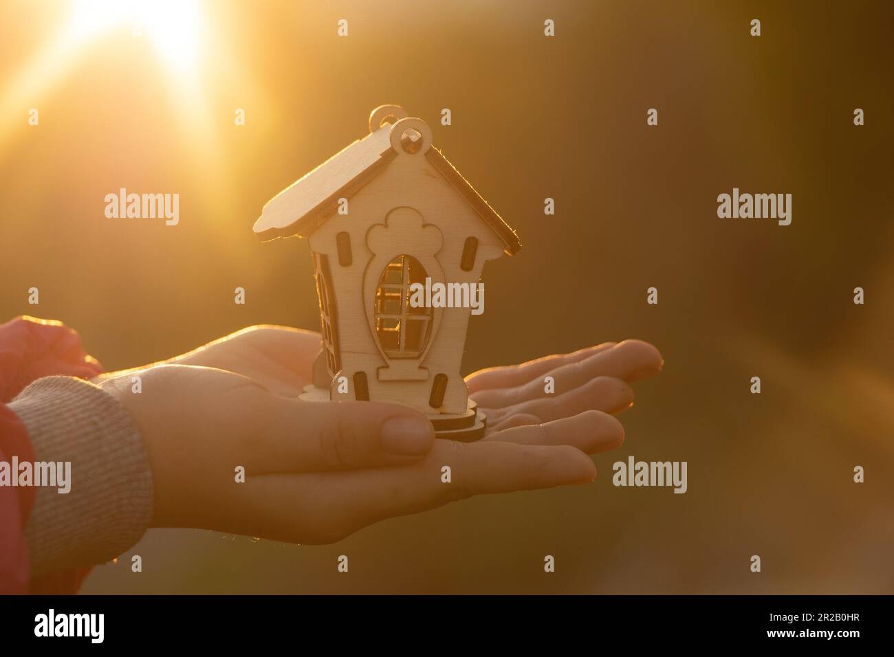 Hände halten ein kleines Holzhaus vor dem Hintergrund der Sonne bei Sonnenuntergang, Traumhaus Stockfoto