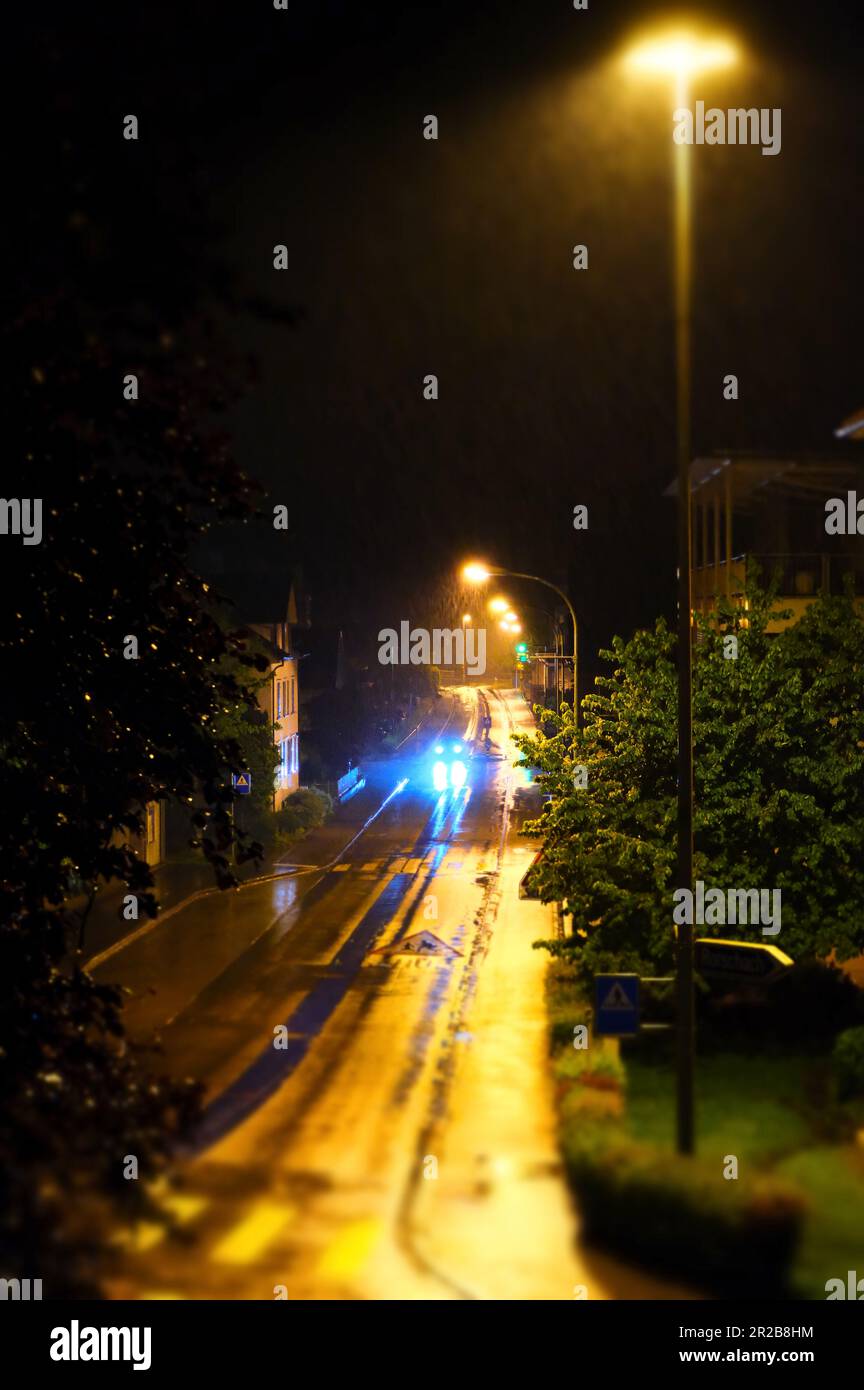 In der stummen Umarmung der Nacht durchquert ein einsames Auto die schwach beleuchtete Straße. Seine Scheinwerfer strahlen ein ätherisches Licht aus und malen die Umgebung. Stockfoto