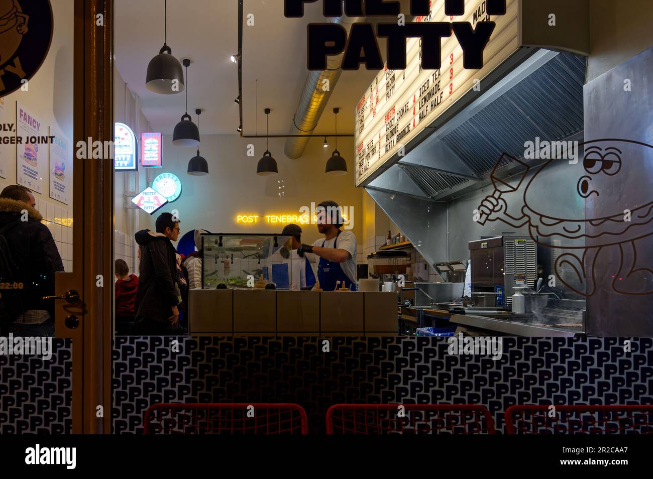 Leute, die in einem kleinen Hamburger-Laden bestellen. Von außen betrachtet man den Zubereitungsbereich eines Fast-Food-Restaurants. Schweizer Burgerladen. Stockfoto