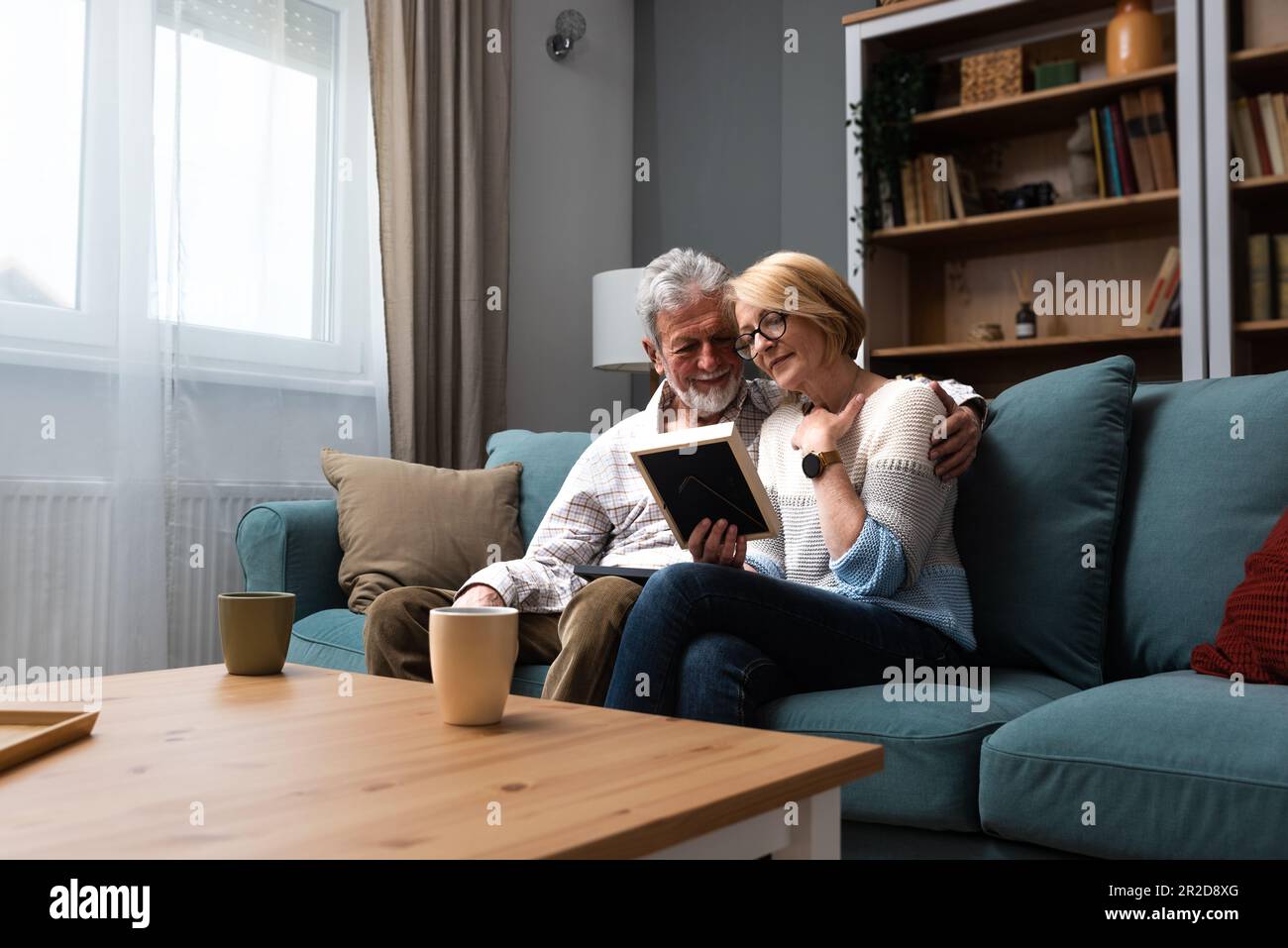 Zwei ältere Leute, ein älteres Paar, Mann und Frau, die ein Familienfoto ansehen, lächeln und umarmen, während sie zu Hause auf dem Sofa sitzen. Erinnerungen und Nostalgie für das Kind Stockfoto
