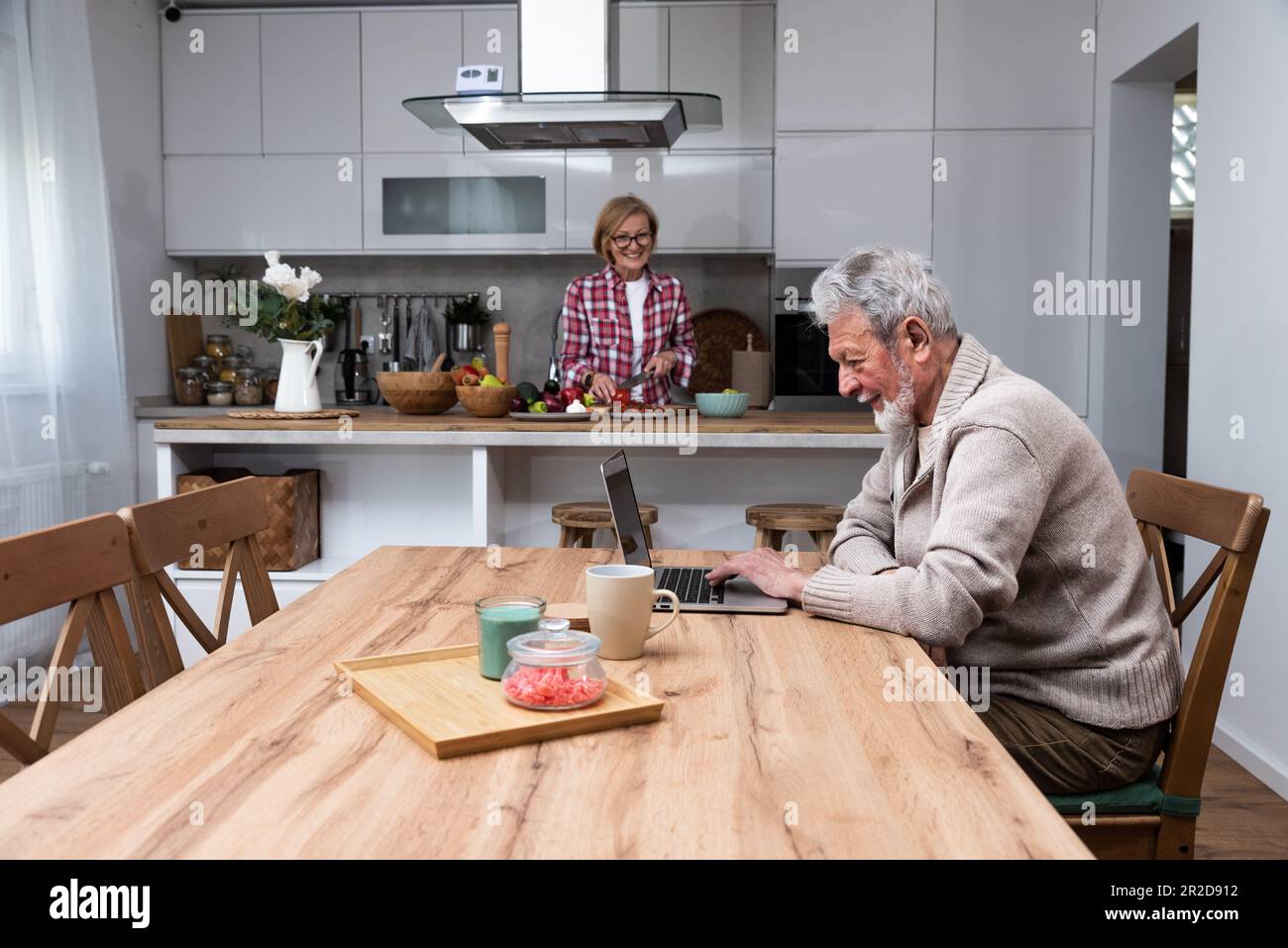 Glücklicher älterer Mann Ehemann, der aus der Ferne arbeitet oder online Fernunterricht mit einem Laptop vom Heimbüro aus lernt, während ihre ältere Frau Frühstück kocht Stockfoto