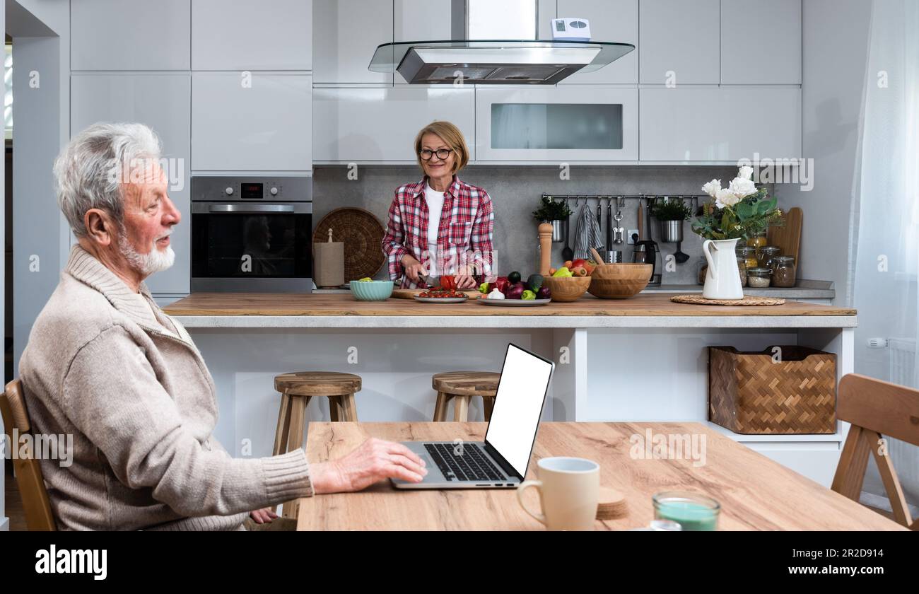 Glücklicher älterer Mann Ehemann, der aus der Ferne arbeitet oder online Fernunterricht mit einem Laptop vom Heimbüro aus lernt, während ihre ältere Frau Frühstück kocht Stockfoto