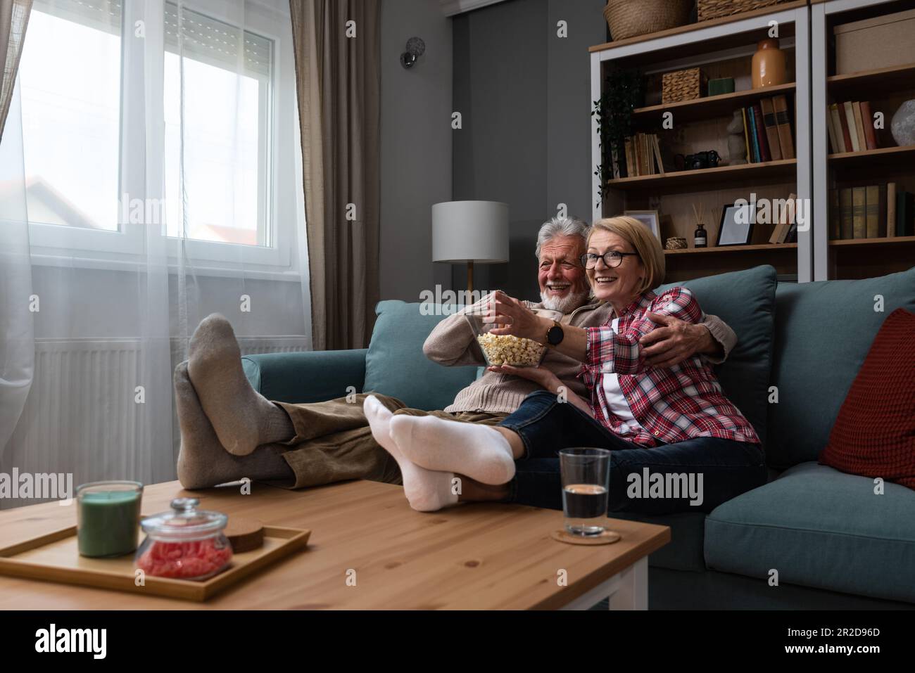 Lachendes älteres Paar, Mann und Frau, die fernsehen, Comedy-Show oder Film schauen und Popcorn-Snack essen, zu Hause auf einer gemütlichen Couch sitzen, ältere Familie, Mann und Stockfoto