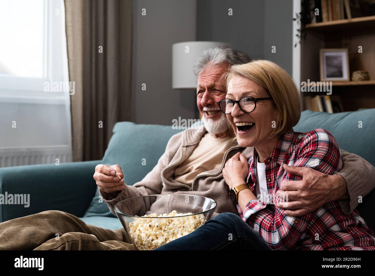 Lachendes älteres Paar, Mann und Frau, die fernsehen, Comedy-Show oder Film schauen und Popcorn-Snack essen, zu Hause auf einer gemütlichen Couch sitzen, ältere Familie, Mann und Stockfoto