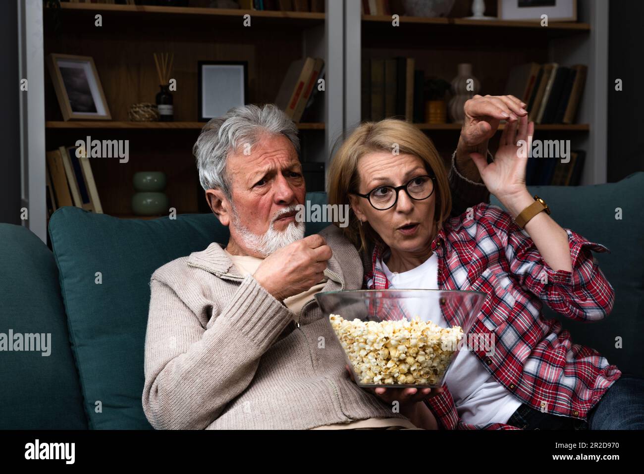 Ein reifes Ehepaar, das sich amüsiert und ein verängstigtes Fernsehprogramm ansieht. Senioren schauen zu Hause Horrorfilme mit Popcorn und ängstlichen Gesichtern. Älter Stockfoto